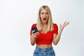 Angry and frustrated blond girl screams, holds mobile phone and shouts emotional, stands against white background