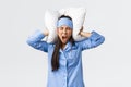 Angry furious asian girl in pajamas and sleeping mask, lying bed and shut ears with pillow, shouting mad as cant sleep
