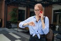 Angry irritated young business woman holding mobile phone and looks at his watch at the blurry business center building Royalty Free Stock Photo