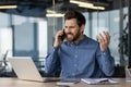 Angry and irritated young businessman sitting in the office at the desk and talking on the phone while waving his hands Royalty Free Stock Photo