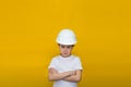 Angry little girl in a construction white helmet crossed her arms over her chest on a yellow background