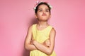 An angry little girl wearing yellow dress standing with crossed arms posing over pink background. Displeased preschooler child