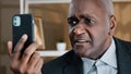 Angry mature elderly African business man having emotionally unpleasant conversation video call with smartphone upset Royalty Free Stock Photo