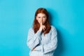 Angry redhead girl hushing at you, show taboo gesture, forbid to speak, standing over blue background in sweater Royalty Free Stock Photo