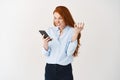 Angry redhead girlfriend looking pissed-off at smartphone screen and grimacing outraged, squeeze hand of rage and