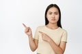 Angry sulking asian girl pointing fingers left, frowning displeased, complaining at smth, white background