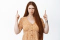 Angry sulking redhead girl pointing fingers up, looking with jealous and upset face expression, standing over white