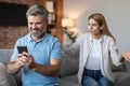 Angry upset adult european wife yelling at smiling husband with smartphone in living room interior