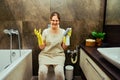 Angry woman sits on the toilet while cleaning the bathroom. Disinfection with yellow rubber gloves in the washroom