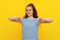 Angry young brunette woman in striped t shirt, looking upset, showing thumbs down, standing over yellow background Royalty Free Stock Photo