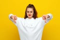 Angry young brunette woman in white casual style sweatshirt, looking upset, showing thumbs down. Indoor studio shot on yellow Royalty Free Stock Photo