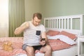 Angry young man working from home in bedroom, holding his phone in hand and a fist raised. Royalty Free Stock Photo