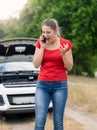 Angry young woman standing at broken car in field and shouting in mobile phone Royalty Free Stock Photo