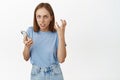 Annoyed blond girl holding her mobile phone, clench fist in anger, staring furious and irritated, bothered by someone Royalty Free Stock Photo