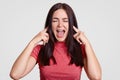 Annoyed brunette woman asks to stop this sound, pluggs ears with both index fingers, exclaims angrily, keeps eyes shut, dressed ca Royalty Free Stock Photo