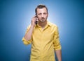 Annoyed handsome guy on the phone over blue background, dresses in yellow shirt. Annoying man talking on smartphone. Nuisance and