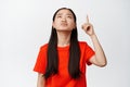 Annoyed teen asian girl pointing and looking up with tired, fed up face expression, being pissed off, standing in red t Royalty Free Stock Photo