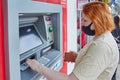 Young white woman wearing PPE gets cash in form of paper currency at street ATM.