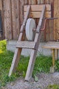 An antique grindstone standing against a wooden shed with overgrown grass. Old grey sharpening stone tool or equipment Royalty Free Stock Photo