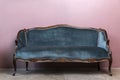 An antique sofa with a wooden base and a blue-green velor seat sits against a pink wall. Front view Royalty Free Stock Photo