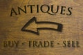 Antiques Buy Trade Sell sign set on a wood background and a left facing arrow. Brown 3D text against wood grain background.