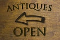 Antiques Open sign set on a wood background and a left facing arrow. Brown 3D text against wood grain background.