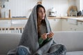 Anxious sick woman sitting covered in blanket holding thermometer, having high body temperature