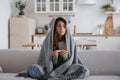 Anxious sick woman sitting covered in blanket holding thermometer, having high body temperature