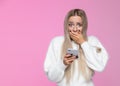 Anxious young blond woman covers mouth in shock, holding mobile phone, looking at camera, surprise reaction on her face, isolated