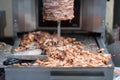 Machine for cooking shawarma, meat layers strung on a skewer, chopped meat is on grill surface, tongs and knife, front view