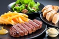 Appetising steak slices served with fries , green salad and baguette on a black plate.