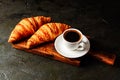 Appetizing breakfast, croissants and coffee in white cup with a spoon and a saucer on a wooden plate, on a dark gray table under Royalty Free Stock Photo