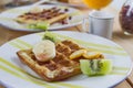 Appetizing fried golden Belgian waffle with pieces of fresh banana, kiwi, kumquat and maple syrup on a striped earthenware plate. Royalty Free Stock Photo
