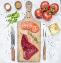 Appetizing raw beef steak with tomatoes, knife and fork for meat on a vintage chopping board, around laid out ingredients, seasoni