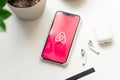 Apple Iphone 11 Pro with red Airbnb logo on display, Airpods with case, credit bank card, plant on white table. Top view
