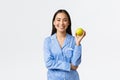 An apple keeps doctors away. Cheerful cute asian girl in blue pajamas eating apple for breakfast and smiling joyful