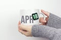 April 18th. Day 18 of month, Calendar date. Closeup of female hands in grey sweater holding cup of tea with month and calendar