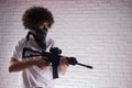 an Arab with an African hairstyle in stylish fashionable clothes with an airsoft gun poses against a brick wall