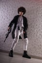 an Arab with an African hairstyle in stylish fashionable clothes with an airsoft gun poses against a brick wall