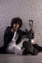 an Arab with an African hairstyle in stylish fashionable clothes with an airsoft gun poses against a brick wall