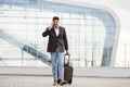 Arab businessman traveller talking on phone, while walking outside modern airport, carrying suitcase Royalty Free Stock Photo