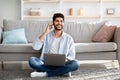 Arab freelancer man using laptop and talking on cellphone, working while sitting on floor in living room at home Royalty Free Stock Photo