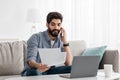 Arab freelancer man working remotely with smartphone, laptop and documents, sitting on sofa at home Royalty Free Stock Photo