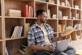 Arab man learning online, reading book and using laptop computer, sitting on floor an leaning on book shelf at home Royalty Free Stock Photo