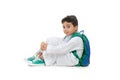 Arab school boy sitting on ground with a smile on his face, wearing white traditional Saudi Thobe, back pack and sneakers, raising