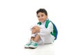 Arab school boy sitting on ground with a smile on his face, wearing white traditional Saudi Thobe, back pack and sneakers, raising