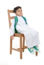 Arab school boy sitting sleepy and tired on a wooden chair, wearing white traditional Saudi Thobe, back pack and sneakers, raising