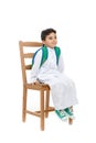 Arab school boy sitting on a wooden chair with a smile on his face, wearing white traditional Saudi Thobe, back pack and sneakers