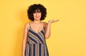 Arab woman with curly hair wearing striped colorful dress over isolated yellow background smiling cheerful presenting and pointing Royalty Free Stock Photo