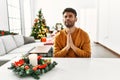 Arab young man sitting on the table by christmas tree begging and praying with hands together with hope expression on face very Royalty Free Stock Photo